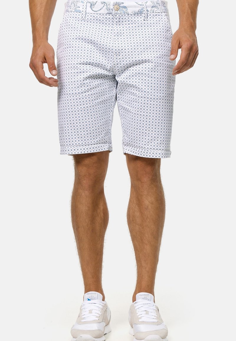Shorts blancs avec un motif circulaire bleu, fabriqués en tissu léger, dotés d'une taille plate et d'ourlets retroussés. Portés avec des baskets blanches.
