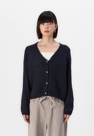 Navyblauer Strickcardigan mit V-Ausschnitt, langen Ärmeln und Perlenknöpfen vorne. Über einem beigen Oberteil getragen und mit hellen Hosen kombiniert.
