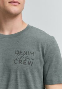 Mees, kes kannab heledat halli ümarat kaelusega T-särki, mille vasakul rinnal on kiri "DENIM Urban CREW".