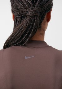 Brun atletisk tröja med korta ärmar, slät textur och en liten grå Nike-logotyp nära halslinningen, med synliga sömnadsdetaljer.