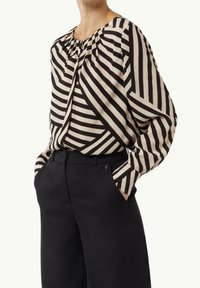 Blusa a maniche lunghe con motivo a righe nere e beige. Presenta una scollatura rotonda con leggere pieghe. Abbinata a pantaloni neri a vita alta.