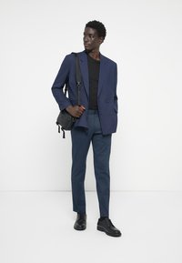 Blazer bleu marine croisé sur un t-shirt noir, assorti à un pantalon bleu texturé et des chaussures noires. Sac bandoulière noir compris.