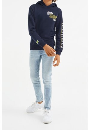 Młodzież w granatowej bluzie z kapturem z napisem "Podążaj za świecącym światłem", w jasnoniebieskich dżinsach i białych trampkach, stojąca z lekko zgiętymi ramionami.