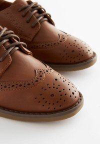 Brogues en cuir marron avec des détails perforés, bout arrondi et lacets marron, présentant une texture lisse et une semelle en caoutchouc basse.