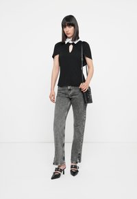 Frau mit schwarzem Oberteil mit weißem Kragen, grauen Acid-Wash-Jeans, schwarzen High Heels und einer schwarzen Umhängetasche, die vor einem weißen Hintergrund steht.