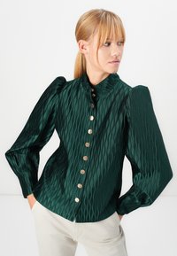 Woman with blonde hair wearing a dark green textured blouse with puffed sleeves and gold buttons, paired with light-colored pants, hands in pockets.