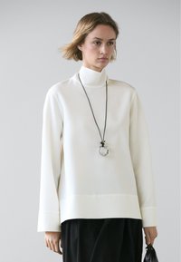 White turtleneck top with long sleeves, features a smooth texture. Paired with a black pendant necklace and black trousers.
