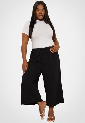 Frau mit langen dunklen Haaren, die ein weißes kurzärmeliges Oberteil, schwarze weit geschnittene Hosen und weiße Sandalen trägt, vor einem einfarbigen Hintergrund stehend.