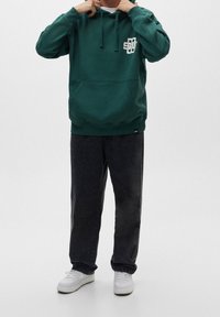 Sudadera verde con un bolsillo tipo canguro, capucha ajustable con cordón y logo blanco en el pecho; combinada con jeans oscuros y zapatillas blancas.