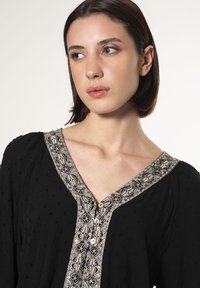 Schwarze Bluse aus strukturiertem Stoff, mit V-Ausschnitt, besticktem Kragen und Knopfverschluss. Verfügt über kleine Akzente und ein lockeres Fit-Design.