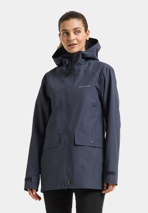 Woman wearing a navy hooded waterproof jacket with front pockets, zip and snap closures, posing against a plain light background.