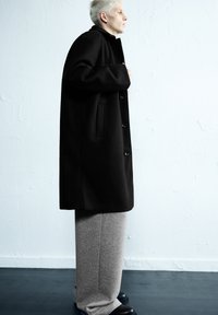 Zwarte wollen jas met hoge kraag, voorzien van een knoopsluiting aan de voorkant en een zijzak. Gecombineerd met grijze joggingbroek en zwarte schoenen.