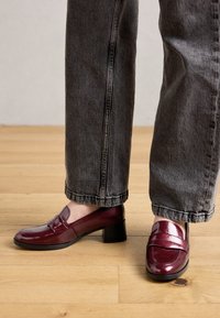Person wearing dark gray jeans and shiny burgundy loafers with low block heels standing on wooden floor.