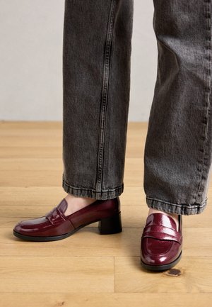 Personne portant un jean gris foncé et des mocassins bordeaux brillants avec de petits talons carrés, se tenant sur un sol en bois.