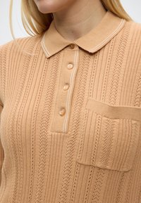 Polo en maille beige avec motifs texturés verticaux, quatre boutons sur la patte de boutonnage et une petite poche sur la poitrine gauche.
