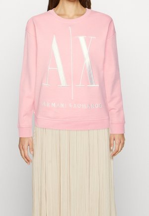 Persoon draagt een roze Armani Exchange sweater met grote witte "AX" letters en een beige geplooide rok tegen een effen achtergrond.