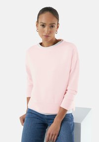 Lichtroze sweatshirt met een ronde halslijn en lange mouwen, gecombineerd met blauwe jeans. Zachte textuur, relaxte pasvorm en zichtbare naaddetails.