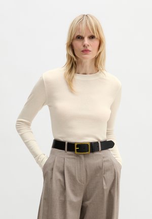 Blonde Frau, die ein cremefarbenes Langarmoberteil trägt, das in beige plissierte Hosen mit einem schwarzen Gürtel und goldener Schnalle gesteckt ist, Hände in den Taschen.