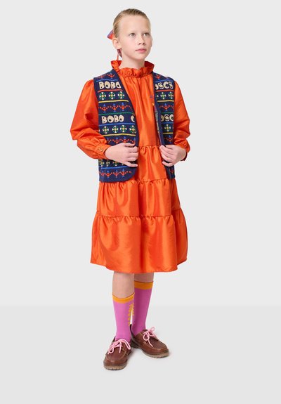 Orange sidentopp med volangkrage och flerlagad kjol, tillsammans med en marinblå väst med färgglada mönster och text. Rosa strumpor och bruna skor.