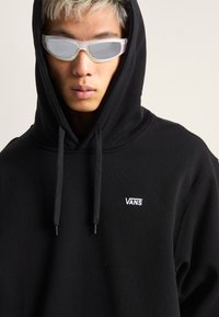 Felpa con cappuccio nera con il logo bianco "VANS" sul petto, dotata di laccetti. Il modello indossa occhiali iridescenti.