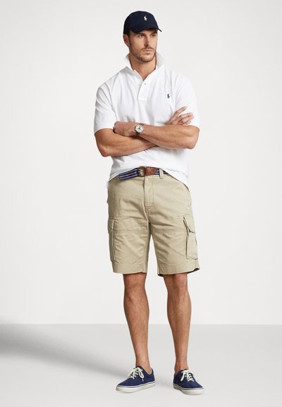 Biała koszulka polo, beżowe krótkie spodenki cargo z bocznymi kieszeniami, pasek w niebieskie paski, granatowa czapka i niebieskie trampki. Klasyczny, casualowy styl.