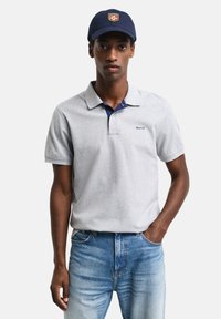 Grå polo skjorte med marineblå krage, har en liten blå logo. Kombinert med lyseblå jeans. Modell med en marineblå caps med logo.