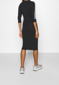 Mujer con un vestido midi negro de manga tres cuartos y rayas blancas, combinado con zapatillas blancas y plateadas, de pie frente a una pared blanca.