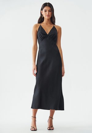 Abito halter in raso nero con profondo scollo a V, corpino aderente e orlo fluente. Sandali con cinturini completano il look.