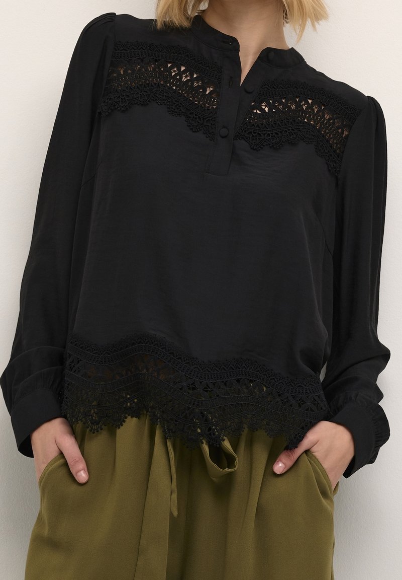 Blouse noire à manches longues avec des détails en dentelle sur la poitrine et l'ourlet, dotée d'un col boutonné. Assortie à un pantalon vert olive.
