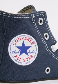 Námořnicky modrá plátěná kotníková teniska s bílou gumovou podrážkou, se znakem "Converse All Star" v modro-červeném kruhu a bílými očky na tkaničky.