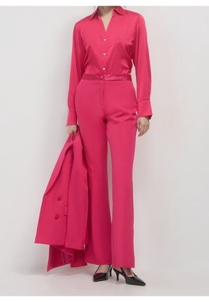 Femme en pantalon et chemise rose vif tenant un blazer assorti, portant des talons pointus noirs, debout devant un fond clair uni.