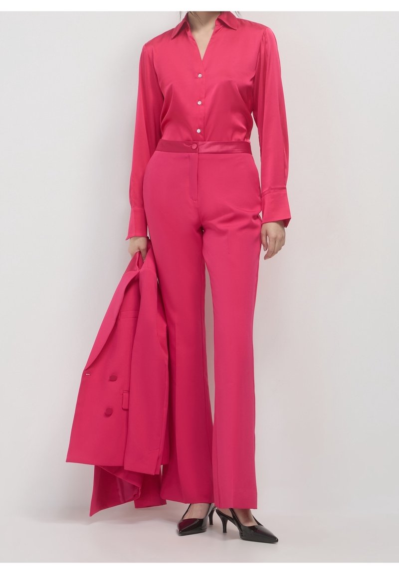 Femme en pantalon et chemise rose vif tenant un blazer assorti, portant des talons pointus noirs, debout devant un fond clair uni.