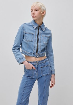 Person mit kurzen blonden Haaren, die eine hellblaue kurze Jeansjacke und hoch taillierte Jeanshose mit großen Fronttaschen trägt.