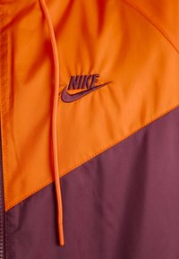 Chaqueta de Nike con diseño en bloques de color en naranja brillante y púrpura oscuro, con una textura suave y logo bordado. Incluye cordón ajustable.