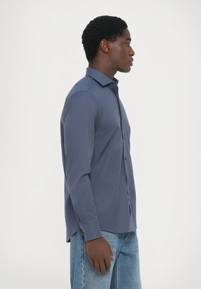 Chemise ajustée à manches longues en tissu bleu marine, avec un col pointu et des boutons, associée à un jean bleu clair. Texture lisse et respirante.