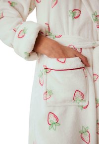 Bata de baño de forro polar blanco con un estampado de fresas. Cuenta con un cinturón de cinta, mangas largas y un bolsillo con ribete rojo. Tejido suave y texturizado.