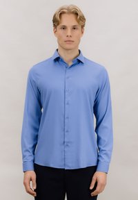 Ung mann med blondt hår iført en lyseblå, knappet skjorte med lange ermer og mørke bukser mot en ensfarget bakgrunn.