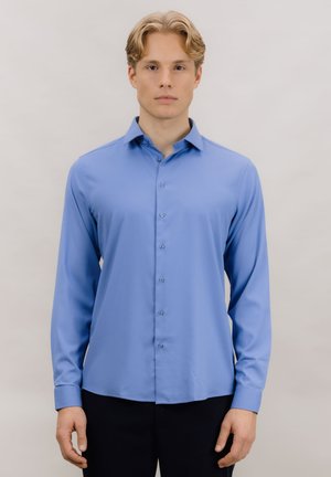 Ung mann med blondt hår iført en lyseblå, knappet skjorte med lange ermer og mørke bukser mot en ensfarget bakgrunn.