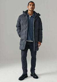 Daunenjacke in Dunkelgrau, mit Kapuze, Reißverschluss und Seitentaschen. Kombiniert mit einem blauen Poloshirt und dunklen Hosen, abgerundet mit schwarzen Stiefeln.