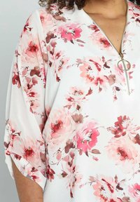 Blusa floreale con fiori rosa e marroni su sfondo bianco, con scollo con zip e maniche a tre quarti.