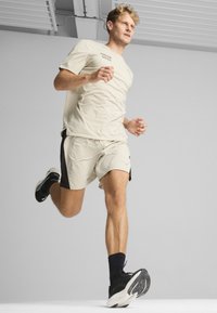 Light beige athletic shirt and shorts with black accents; model running in black sneakers with textured soles, set against a neutral background.