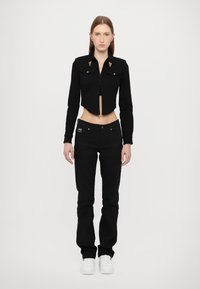 Black cropped jacket with gold hardware, paired with black straight-leg jeans. Model wearing white sneakers. Plain background.