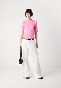 Vrouw staat tegen een witte achtergrond in een roze top, brede witte broek, zwarte riem, houdt een zwarte handtas vast en draagt zwarte puntige schoenen.