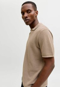 Homme aux cheveux courts et boucle d'oreille en anneau portant un polo à manches courtes beige texturé, de profil sur un fond blanc uni.