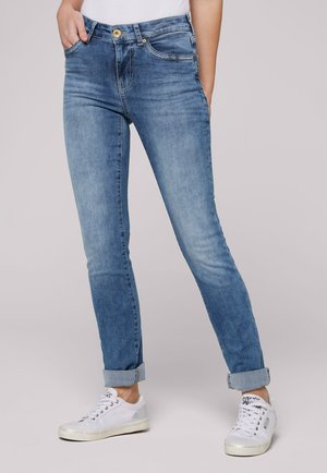 Woman wearing light blue cuffed jeans and white lace-up sneakers, standing with hands in pockets against plain background.