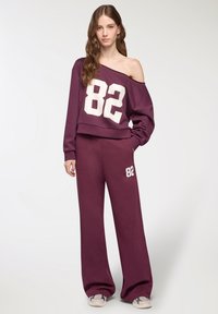 Młoda kobieta ubrana w luźny bordowy dres z białym numerem "82", bluzę odsłaniającą ramię, szerokie spodnie i trampki, pozująca z ręką w kieszeni.