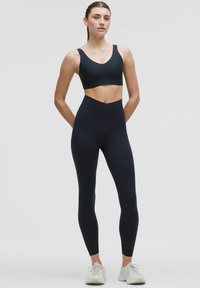 Sujeción deportiva negra y leggings de talle alto hechos de un tejido elástico, con un diseño sin costuras y una textura suave. Zapatillas blancas completan el conjunto.