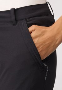 Pantalons noirs avec une poche zippée, une taille plate et une texture lisse. Gros plan sur une main dans la poche et les détails de couture.