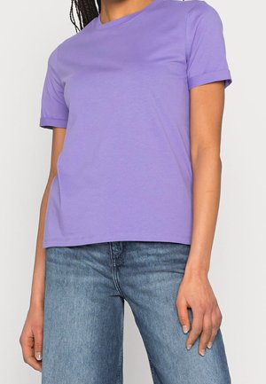 Osoba w gładkiej lawendowej koszulce z krótkim rękawem oraz niebieskich dżinsach o średnim odcieniu, stojąca ze swobodnie opuszczonymi ramionami.