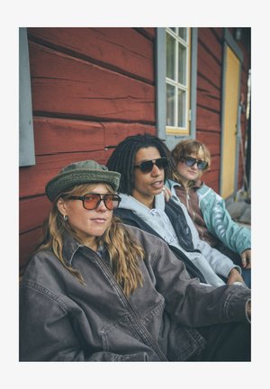 Trois personnes sont assises contre un mur en bois rouge. Elles portent des lunettes de soleil ; l'une porte un chapeau vert et une veste marron, une autre un sweat à capuche gris, et la dernière une veste bleue.